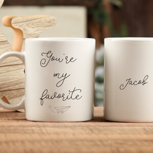 Op de afbeelding: Twee witte keramische mokken met zwarte tekst. De ene mok zegt "You're my favorite" met een papieren vliegtuiggrafiek. De andere mok zegt "Jacob".