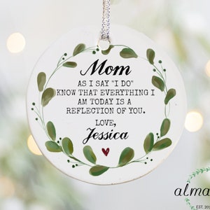 May include: White round ornament with a green leafy wreath and the text "Mom as I say "I do" know that everything I am today is a reflection of you. Love, Jessica"