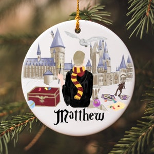 May include: A white ceramic ornament featuring a Hogwarts castle scene with a wizard in a black robe and a red and gold scarf. The ornament has the name "Matthew" written on the bottom.