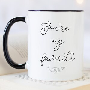Op de afbeelding: Witte keramische mok met een zwarte handgreep en zwarte rand. De mok heeft de tekst "You're my favorite" geschreven in zwarte cursieve letters met een klein papieren vliegtuigje en een hartje.