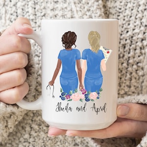 May include: White ceramic mug with a design of two nurses standing back to back. The nurses are wearing blue scrubs and have brown and blonde hair. The mug has a floral design at the bottom and the text "Sheila and April" at the bottom.
