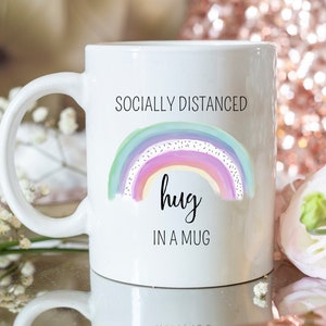 May include: White ceramic mug with a handle, featuring a watercolor rainbow design in pastel colors. The text on the mug reads "Socially Distanced Hug in a Mug". The mug is a standard size for coffee or tea.