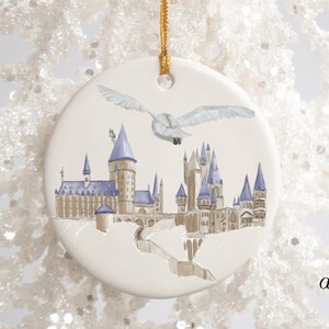 May include: A round, off-white ceramic ornament featuring a watercolor illustration of a castle with blue spires and a flying owl. The ornament has a gold string for hanging. The castle is set against a white background.