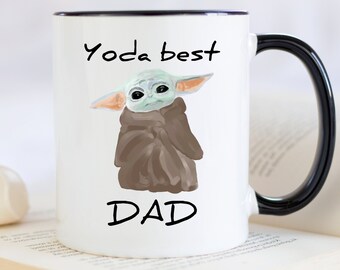 Op de afbeelding: Witte keramische mok met een zwarte rand en handvat. De mok heeft een cartoon afbeelding van een groen buitenaards wezen met de tekst "Yoda best DAD" erop gedrukt.
