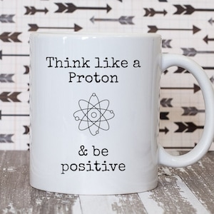 May include: White ceramic mug with a black graphic that says "Think like a Proton & be positive". The graphic also includes a black and white illustration of an atom.