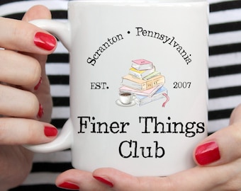 Op de afbeelding: Witte keramische mok met een zwart-wit ontwerp. De mok heeft de tekst "Scranton, Pennsylvania EST. 2007 Finer Things Club" en een tekening van een stapel boeken met een kopje koffie erop.