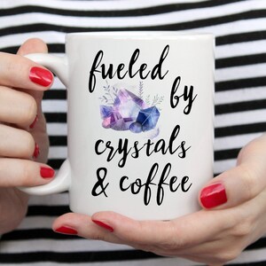 Puede incluir: Taza de cer&aacute;mica blanca con un dise&ntilde;o negro que dice "fueled by crystals & coffee". El dise&ntilde;o tambi&eacute;n incluye una ilustraci&oacute;n de acuarela de cristales morados y azules.