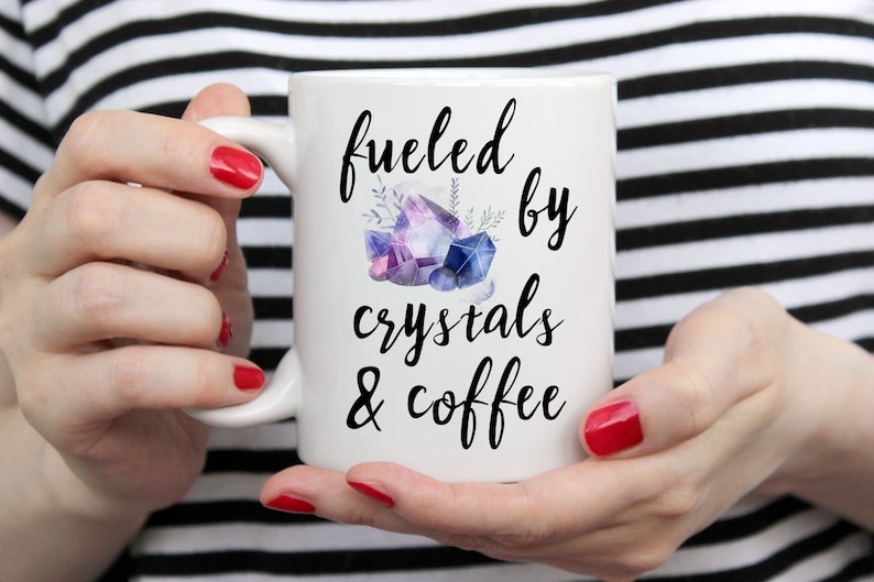 Puede incluir: Taza de cer&aacute;mica blanca con un dise&ntilde;o negro que dice "fueled by crystals & coffee". El dise&ntilde;o tambi&eacute;n incluye una ilustraci&oacute;n de acuarela de cristales morados y azules.