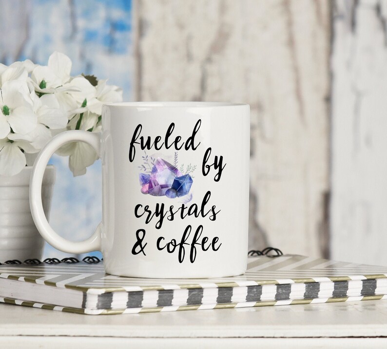 Puede incluir: Taza de cer&aacute;mica blanca con un dise&ntilde;o negro que dice "fueled by crystals & coffee". El dise&ntilde;o tambi&eacute;n incluye una ilustraci&oacute;n de acuarela de cristales morados y azules.