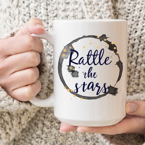Puede incluir: Taza de caf&eacute; de cer&aacute;mica blanca con un anillo de caf&eacute; negro y estrellas doradas. El texto "Rattle the stars" est&aacute; escrito en azul dentro del anillo.