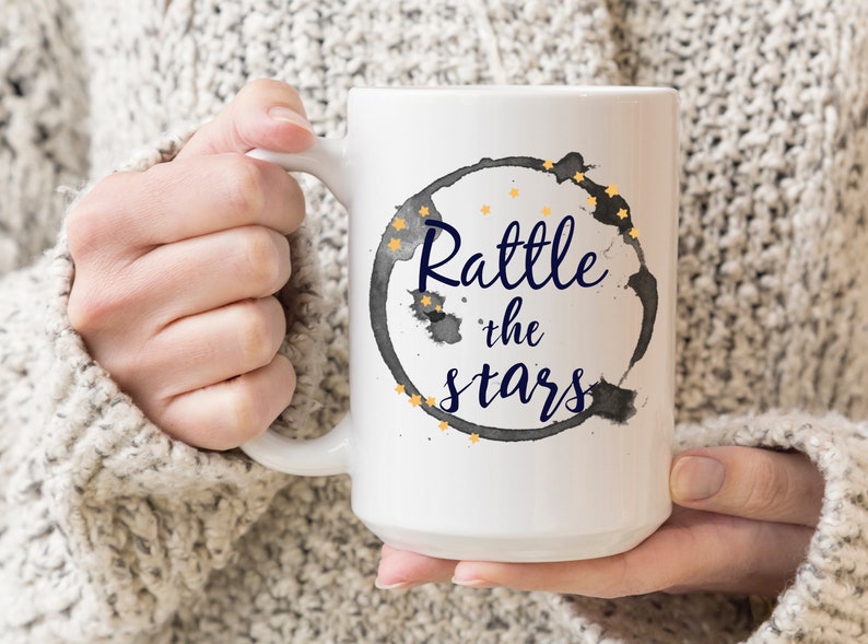 Puede incluir: Taza de caf&eacute; de cer&aacute;mica blanca con un anillo de caf&eacute; negro y estrellas doradas. El texto "Rattle the stars" est&aacute; escrito en azul dentro del anillo.