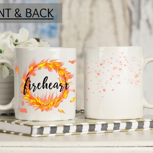 Puede incluir: Taza de cer&aacute;mica blanca con un dise&ntilde;o negro y naranja. El dise&ntilde;o presenta un anillo de fuego con la palabra "fireheart" escrita en su interior. La parte posterior de la taza tiene un dise&ntilde;o naranja y amarillo disperso.
