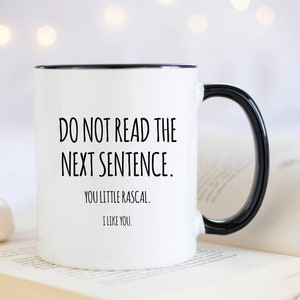May include: White ceramic mug with a black rim and handle. The mug features the text "DO NOT READ THE NEXT SENTENCE. YOU LITTLE RASCAL. I LIKE YOU." in black, centered on the front. The mug is set against a blurred background.