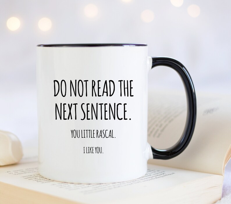 May include: White ceramic mug with a black rim and handle. The mug features the text "DO NOT READ THE NEXT SENTENCE. YOU LITTLE RASCAL. I LIKE YOU." in black, centered on the front. The mug is set against a blurred background.