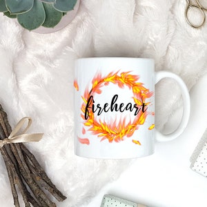Puede incluir: Taza de cer&aacute;mica blanca con un dise&ntilde;o de fuego negro y amarillo y la palabra "fireheart" en letras negras.