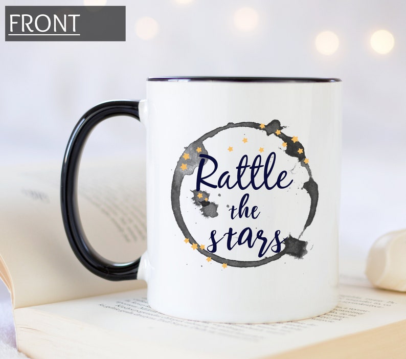 Puede incluir: Taza de cer&aacute;mica blanca con borde y asa negros. La taza presenta un dise&ntilde;o de acuarela en blanco y negro de un c&iacute;rculo con el texto "Rattle the stars" en letras azul marino y estrellas doradas.