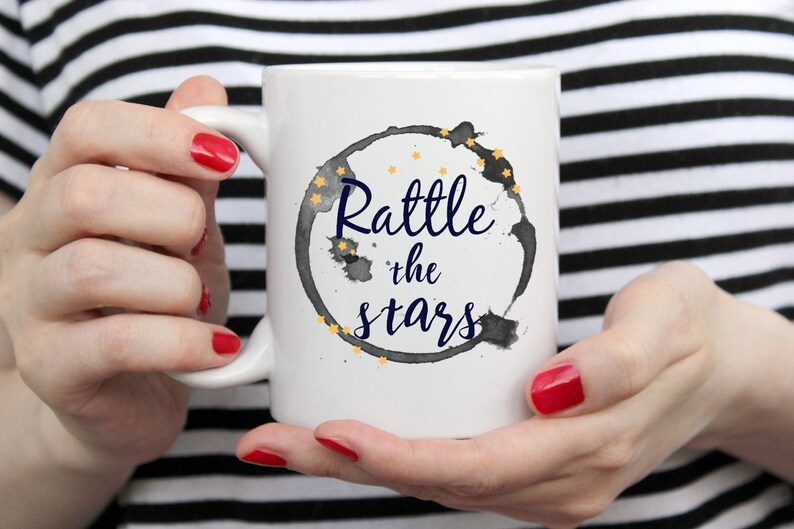 Puede incluir: Taza de caf&eacute; de cer&aacute;mica blanca con un dise&ntilde;o circular de acuarela en negro y gris. El c&iacute;rculo tiene estrellas doradas y el texto "Rattle the stars" en azul marino.