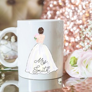 Pu&ograve; includere: Tazza in ceramica bianca con un'illustrazione disegnata a mano di una sposa in abito da sposa bianco che tiene un bouquet di fiori rosa. Il testo "Mrs. Smith" &egrave; scritto sotto la sposa.