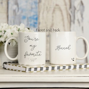 Op de afbeelding: Twee witte keramische mokken met zwarte letters. De ene mok zegt "You're my favorite" met een papieren vliegtuigje. De andere mok zegt "Hannah" met twee kleine hartjes.