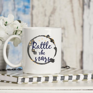 Puede incluir: Taza de cer&aacute;mica blanca con un dise&ntilde;o de acuarela en blanco y negro de un c&iacute;rculo rodeado de estrellas doradas. El texto "Rattle the stars" est&aacute; escrito en azul dentro del c&iacute;rculo.