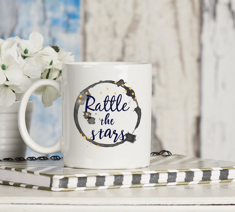 Puede incluir: Taza de cer&aacute;mica blanca con un dise&ntilde;o de acuarela en blanco y negro de un c&iacute;rculo rodeado de estrellas doradas. El texto "Rattle the stars" est&aacute; escrito en azul dentro del c&iacute;rculo.
