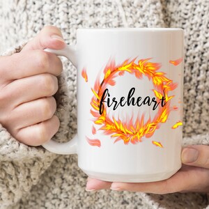 Puede incluir: Taza de cer&aacute;mica blanca con un dise&ntilde;o de un anillo de llamas naranja y amarillo que se arremolina con la palabra "fireheart" en letras negras.
