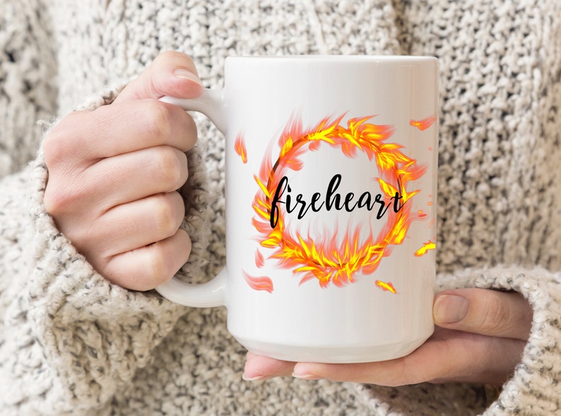 Puede incluir: Taza de cer&aacute;mica blanca con un dise&ntilde;o de un anillo de llamas naranja y amarillo que se arremolina con la palabra "fireheart" en letras negras.