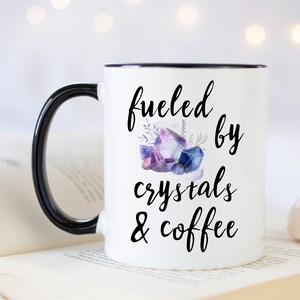 Puede incluir: Taza de cer&aacute;mica blanca con asa y borde negros. La taza tiene un gr&aacute;fico negro que dice "fueled by crystals & coffee" con una ilustraci&oacute;n de acuarela de cristales morados y azules.