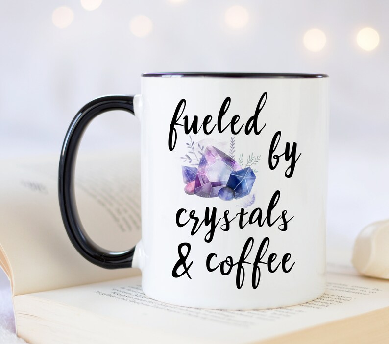 Puede incluir: Taza de cer&aacute;mica blanca con asa y borde negros. La taza tiene un gr&aacute;fico negro que dice "fueled by crystals & coffee" con una ilustraci&oacute;n de acuarela de cristales morados y azules.
