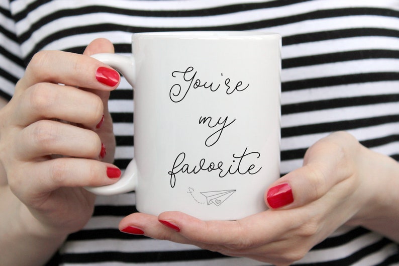 Op de afbeelding: Witte keramische mok met zwarte cursieve tekst die "You're my favorite" luidt en een klein papieren vliegtuigje met een hartje.