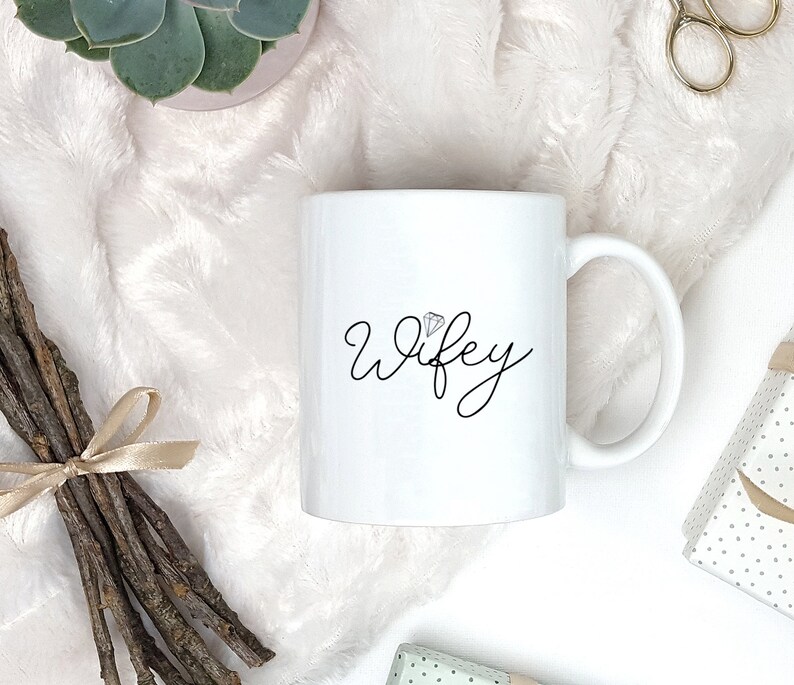 Pu&ograve; includere: Tazza in ceramica bianca con una scritta in corsivo nero che recita "wifey" con un piccolo grafico di diamante sopra la parola.