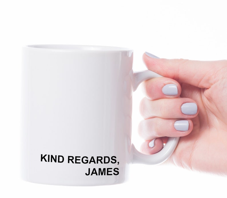 Puede incluir: Taza de cer&aacute;mica blanca con texto negro que dice "KIND REGARDS, JAMES". Una mano sostiene la taza.