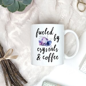 Puede incluir: Taza de caf&eacute; de cer&aacute;mica blanca con texto negro que dice "fueled by crystals & coffee". Una ilustraci&oacute;n de acuarela de cristales morados y azules est&aacute; sobre el texto.