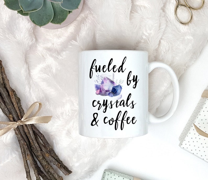 Puede incluir: Taza de caf&eacute; de cer&aacute;mica blanca con texto negro que dice "fueled by crystals & coffee". Una ilustraci&oacute;n de acuarela de cristales morados y azules est&aacute; sobre el texto.