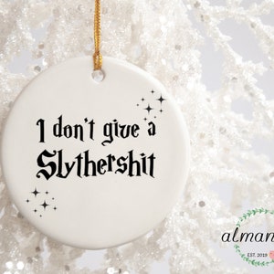 May include: White ceramic Christmas ornament with black text that reads "I don't give a Slythershit" and black star-shaped glitter accents.