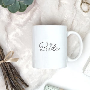 Pu&ograve; includere: Tazza in ceramica bianca con una scritta in corsivo nero che dice "Bride" e un piccolo grafico di diamante sopra la parola.