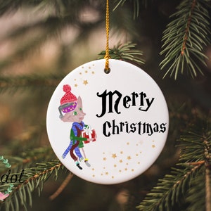 May include: White ceramic Christmas ornament with a cartoon illustration of a cat wearing a red and white hat, green scarf, and holding a wrapped gift. The ornament has gold stars and the text "Merry Christmas" in black.