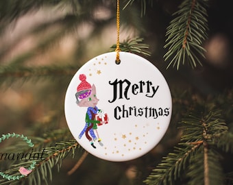 Op de afbeelding: Witte kerstbal van keramiek met een cartoonillustratie van een kat die een rood-witte muts, een groene sjaal draagt en een ingepakt cadeau vasthoudt. De kerstbal heeft gouden sterren en de tekst "Merry Christmas" in zwart.