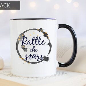Puede incluir: Taza de cer&aacute;mica blanca con borde y asa negros. La taza tiene un c&iacute;rculo de acuarela negro y gris con estrellas doradas y el texto "Rattle the stars" en azul marino.