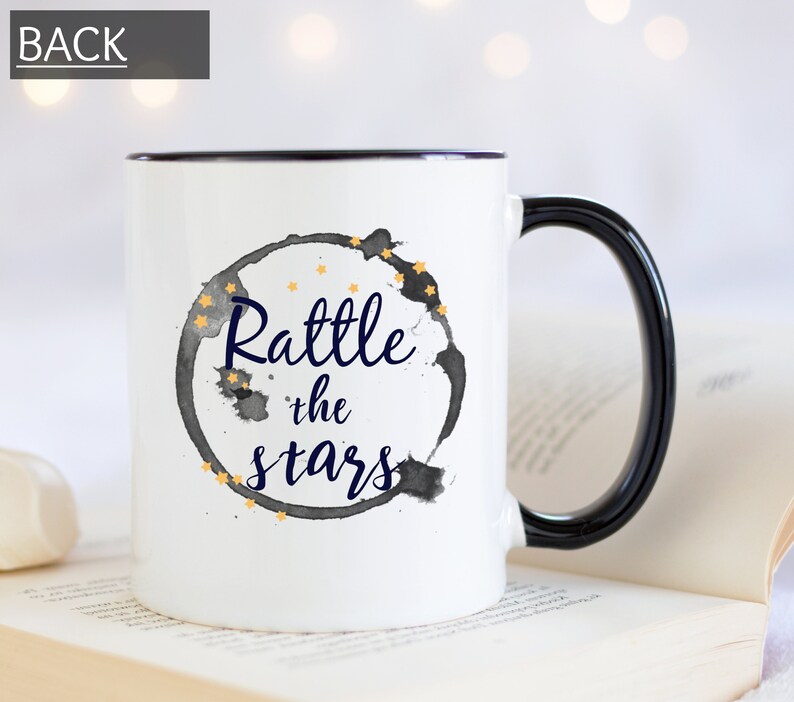 Puede incluir: Taza de cer&aacute;mica blanca con borde y asa negros. La taza tiene un c&iacute;rculo de acuarela negro y gris con estrellas doradas y el texto "Rattle the stars" en azul marino.