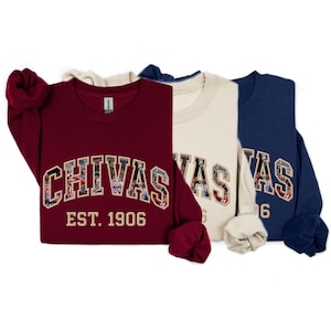 Op de afbeelding: Drie crewneck sweatshirts in bordeaux, crème en marineblauw. Elke sweatshirt heeft het woord "CHIVAS" in een lettertype in collegestijl, met een collage van afbeeldingen in de letters. De tekst "EST. 1906" staat eronder.