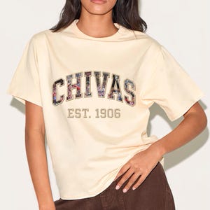 Könnte beinhalten: Ein cremefarbenes Kurzarm-T-Shirt mit dem Wort CHIVAS in großen Blockbuchstaben, die mit einer mehrfarbigen Fotocollage gefüllt sind. Unter dem Text sind die Wörter EST. 1906 in einer einfachen, dunklen serifenlosen Schriftart aufgedruckt.