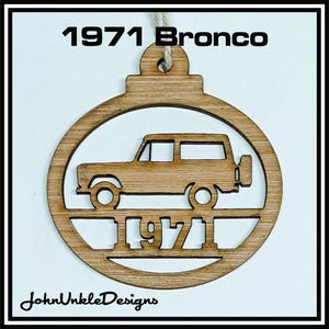 May include: Wooden ornament featuring a cutout of a 1971 Bronco SUV inside a circular frame. The text "1971 Bronco" is at the top, and "1971" is at the bottom. The ornament is made of light brown wood and has a string for hanging. The text "JohnUnkleDesigns" is at the bottom.