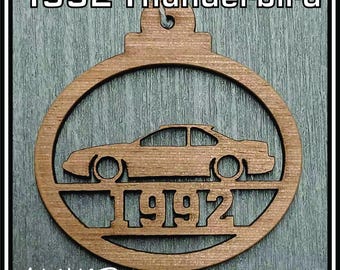 May include: A circular brown wooden ornament featuring a laser-cut silhouette of a 1992 Thunderbird car. The text 1992 is cut out below the car. The ornament has a small loop at the top for hanging. The design is flat with a visible wood grain texture.