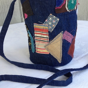 May include: A denim bucket bag with a patchwork design. The bag features a long, thin strap and is adorned with various fabric patches in shades of brown, red, blue, and tan. The interior lining is a light blue fabric.