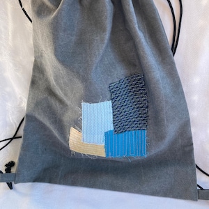 May include: A gray drawstring bag with a patchwork design on the front. The patchwork is made of blue, light blue, and beige fabric.