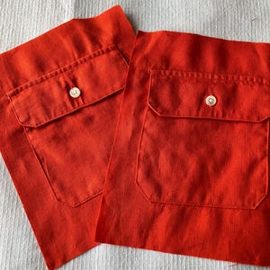 May include: Two pieces of bright orange fabric with attached pockets and a button closure. The fabric has a textured appearance, and the pockets are rectangular with rounded corners. The buttons are white with a subtle, pearlescent sheen.