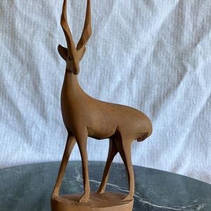 May include: A light brown wooden sculpture of a standing gazelle with long, slender legs and tall, pointed, faceted horns. The animal is depicted in a profile stance, featuring smooth, carved surfaces and a base that supports its four legs.