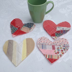 May include: Four patchwork heart-shaped coasters in red, pink, and white fabric with a green ceramic mug on a white tablecloth.