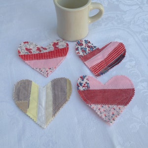 May include: Four handmade patchwork heart-shaped coasters in pink, red, white, and brown fabric. The coasters are sitting on a white tablecloth next to a white ceramic mug.